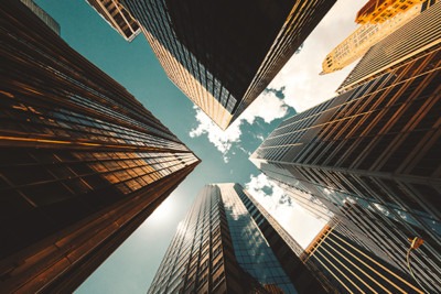 Looking up at towering skyscrapers with reflective glass facades, framed by a bright blue sky and scattered clouds, showcasing urban architecture.