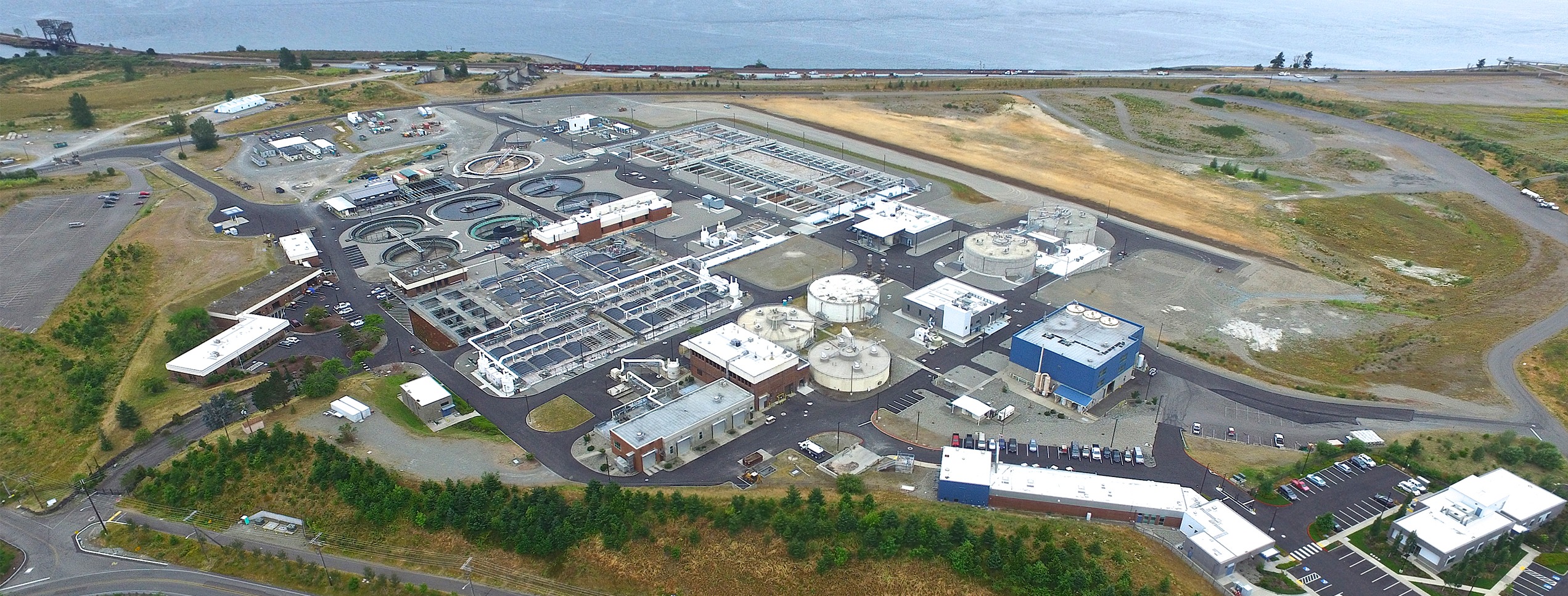 Aerial view of a wastewater treatment facility featuring multiple circular tanks, processing buildings, parking, and nearby natural landscape.