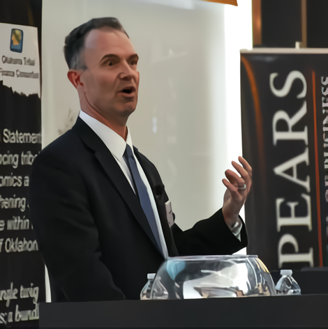 A speaker in a formal suit gestures while presenting at a podium, discussing business topics at a conference. Banners are visible in the background.
