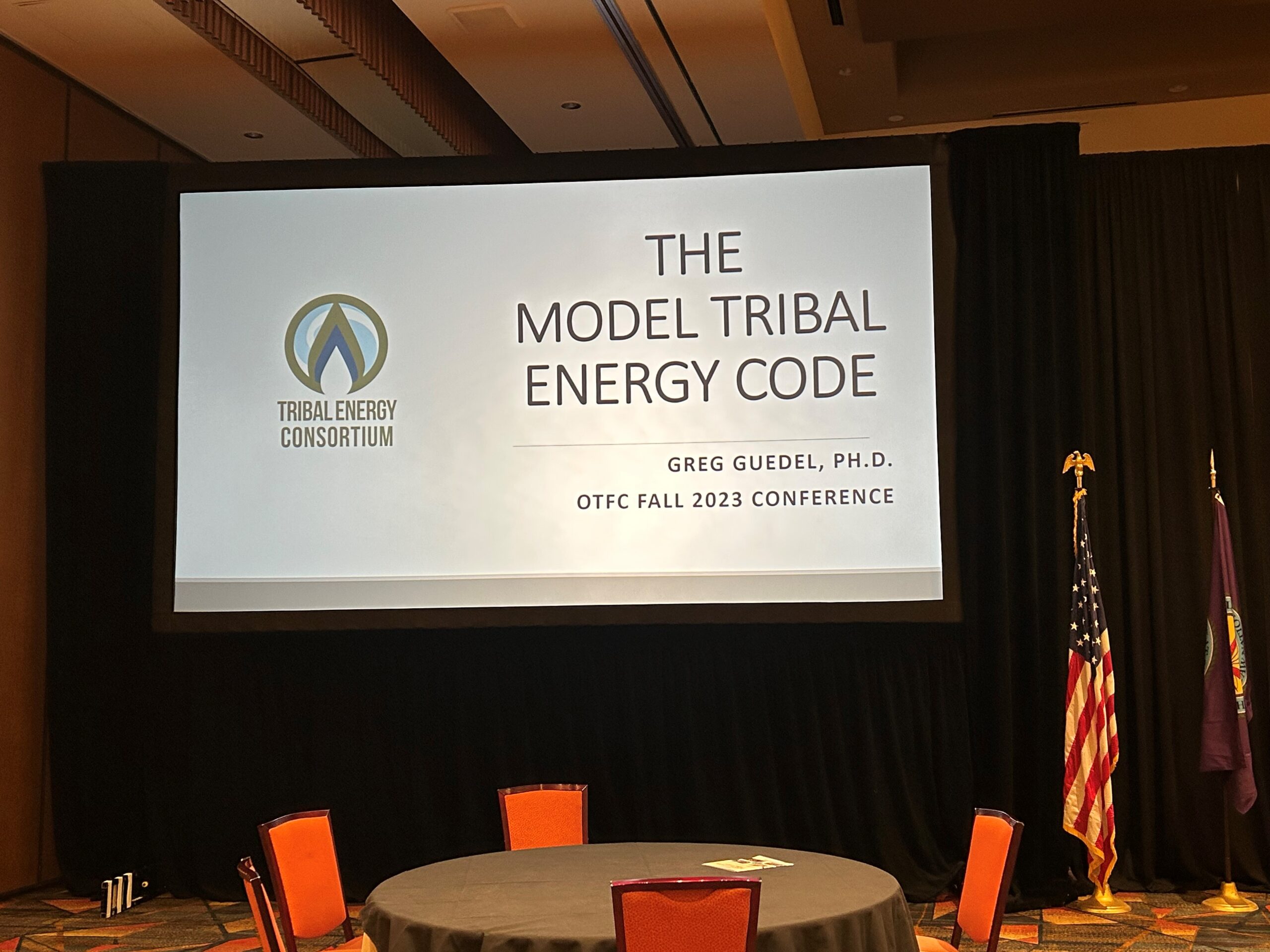Presentation slide titled "The Model Tribal Energy Code" by Greg Guedel, Ph.D., shown at the OTFC Fall 2023 Conference. Red chairs surround a table.